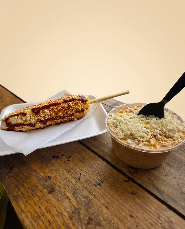 Un elote entero en palito con mayonesa, queso y chile, junto a un vaso de esquites